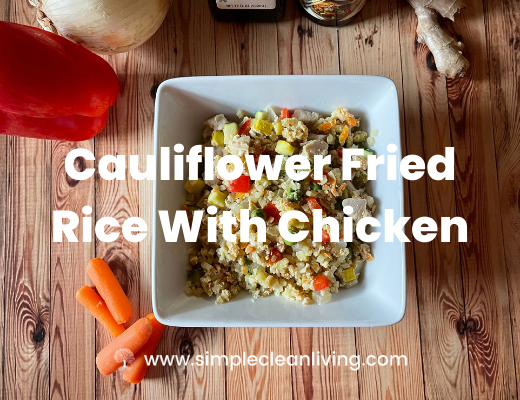 A bowl of cauliflower fried rice with chicken on a table surrounded by the fresh ingredients used to make it