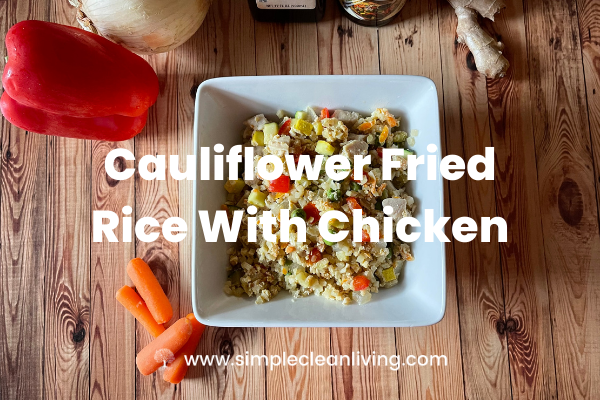 A bowl of cauliflower fried rice with chicken on a table surrounded by the fresh ingredients used to make it