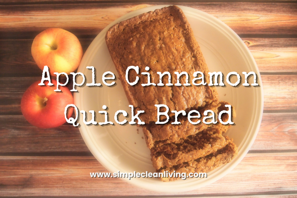 Apple cinnamon quick bread recipe title with picture of a loaf of apple cinnamon bread. The bread is sitting on a white plate on a wooden tabletop. Next to the plate are two fresh apples